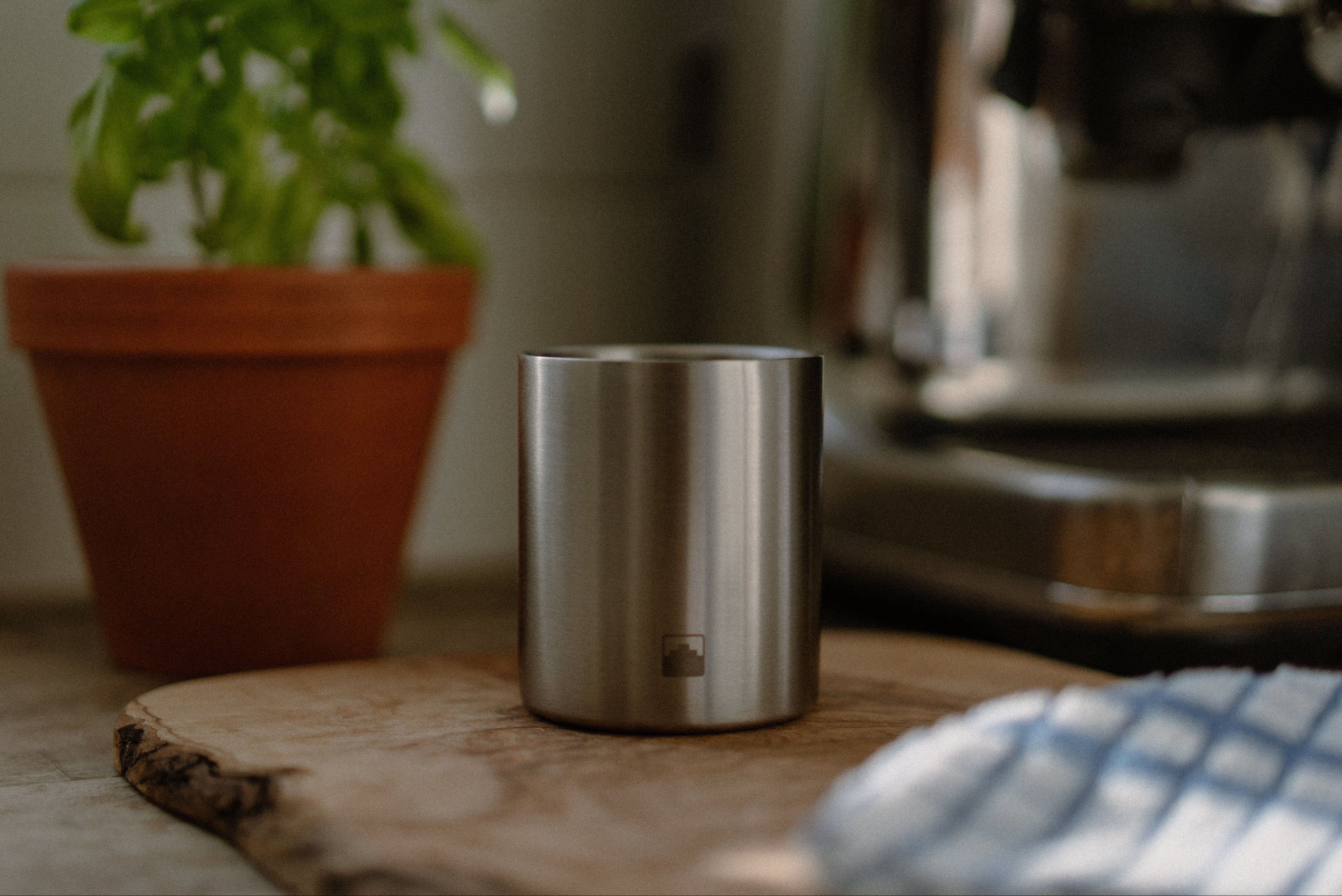 Stainless steel Babel Organic candle on a wooden surface with a blurred plant and coffee machine in the background