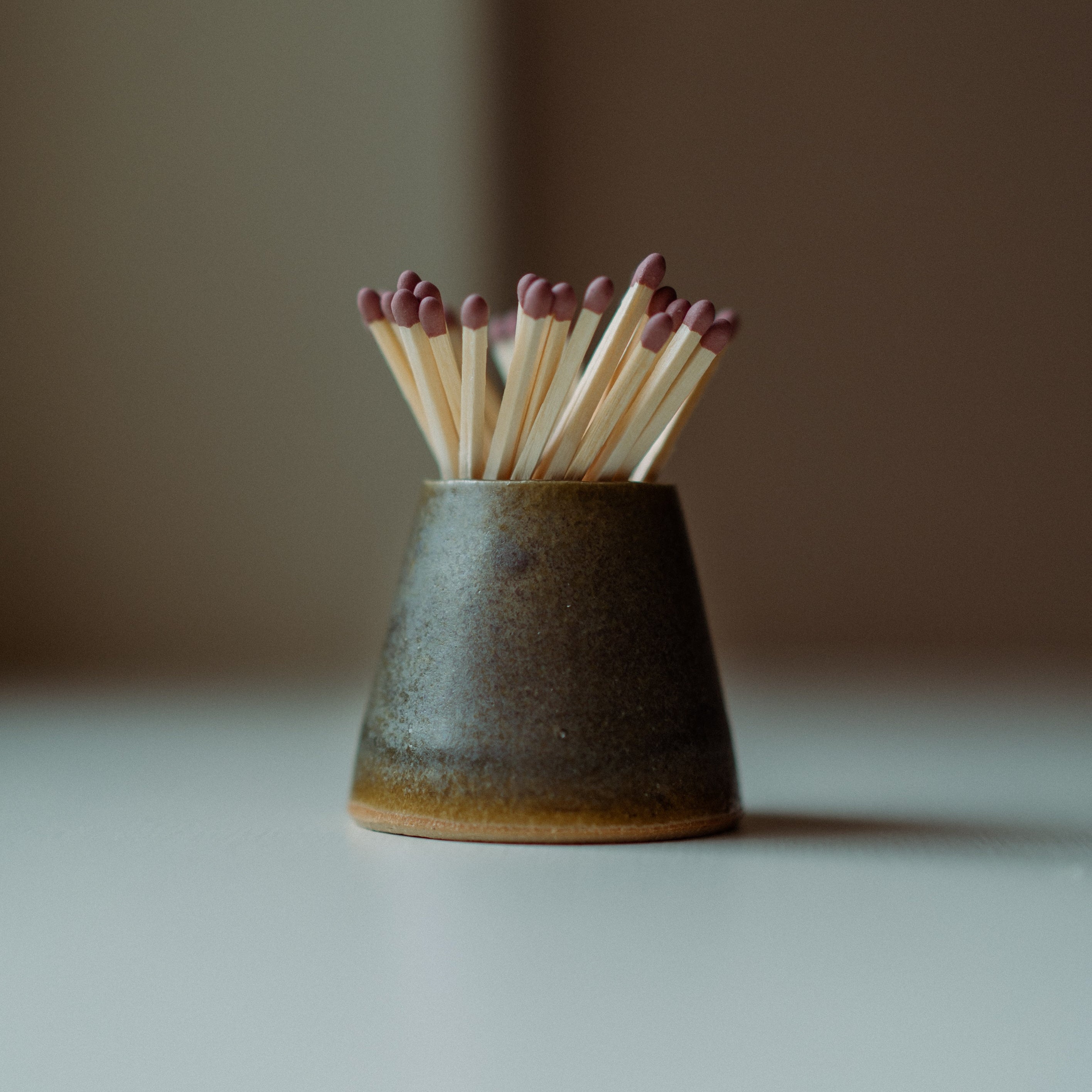 A small dark ceramic match holder on a light surface with wooden matchsticks and reddish tips standing inside it, photographed against a softly blurred neutral background.