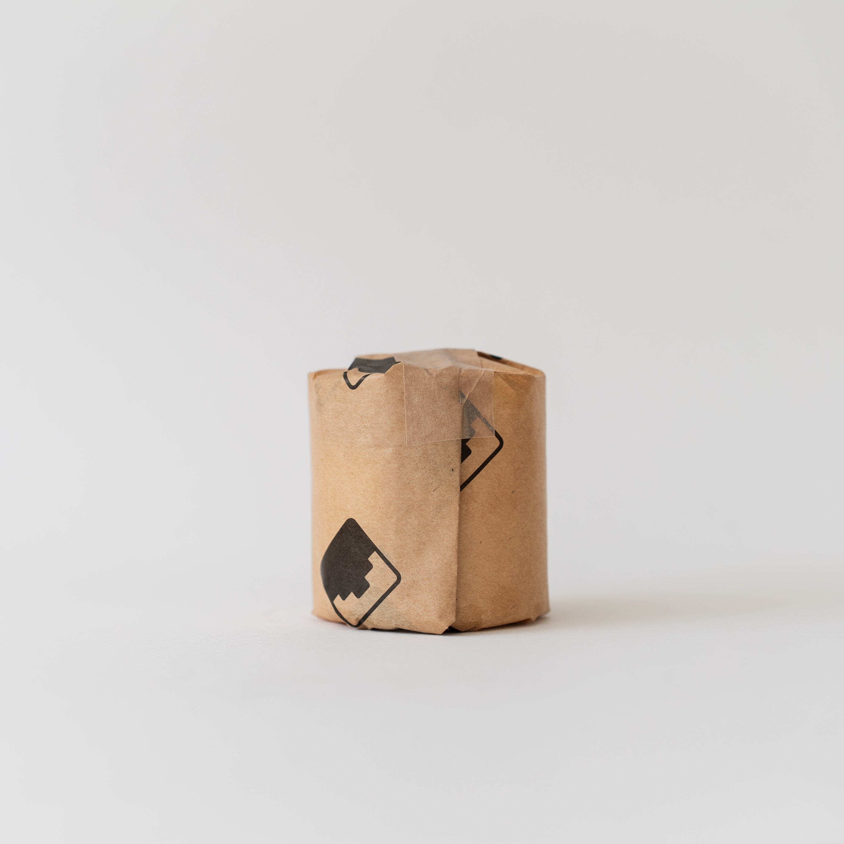 A small cylindrical-shaped candel refill wrapped in light brown kraft paper with black printed Babel logos, standing upright on a seamless light gray background.